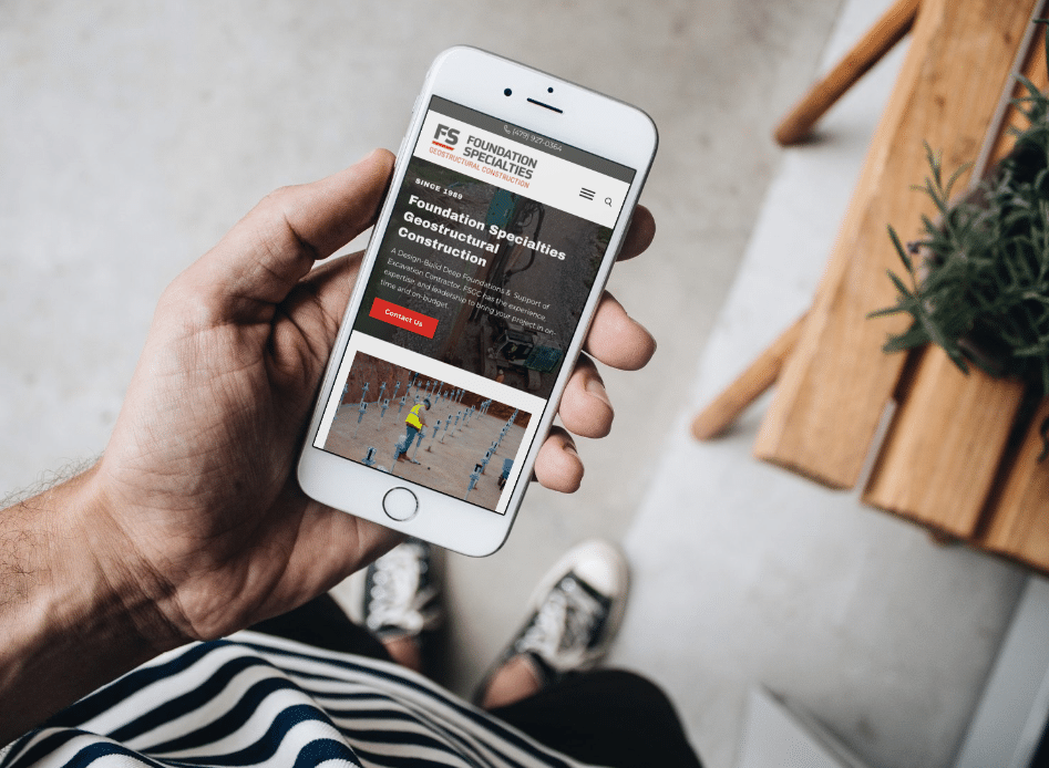 A person holding a white smartphone displaying the Foundation Specialists website, standing indoors near a wooden table with a plant, wearing black and white sneakers and a striped shirt.